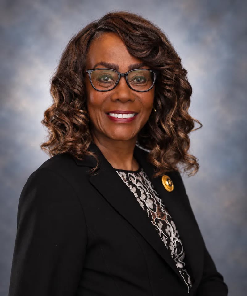 Dianne Oliphant, smiling in glasses and a black blazer against a softly textured blue-grey studio background.