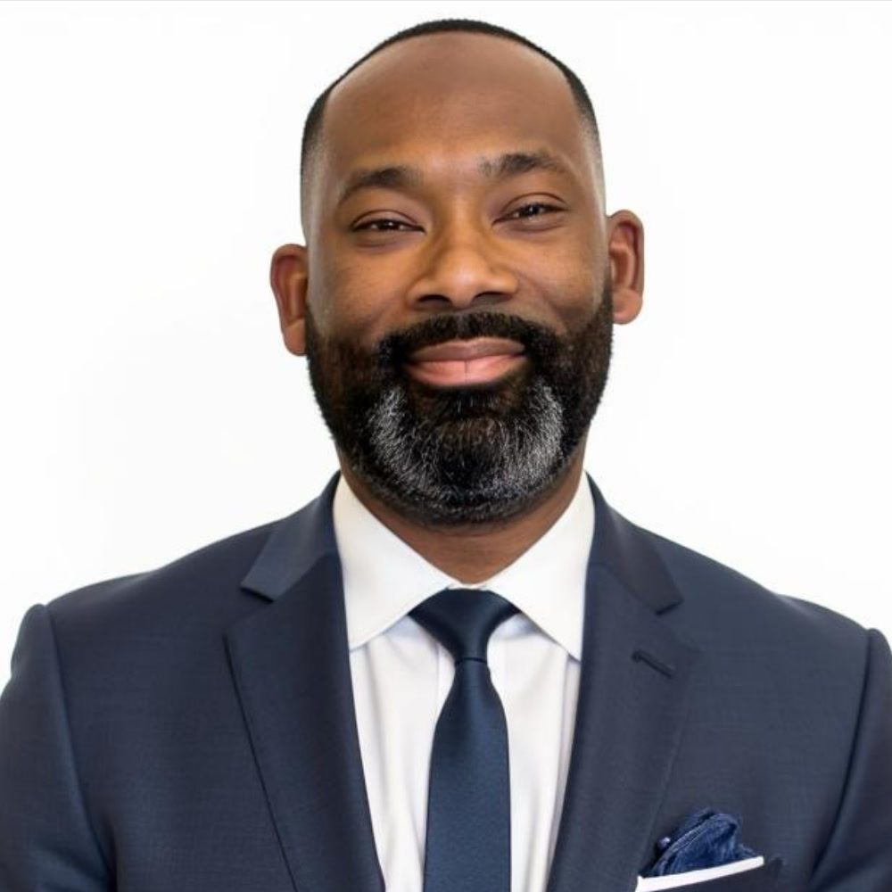 George Reed Jr., in a navy suit and tie, smiling against a dark grey studio background.
