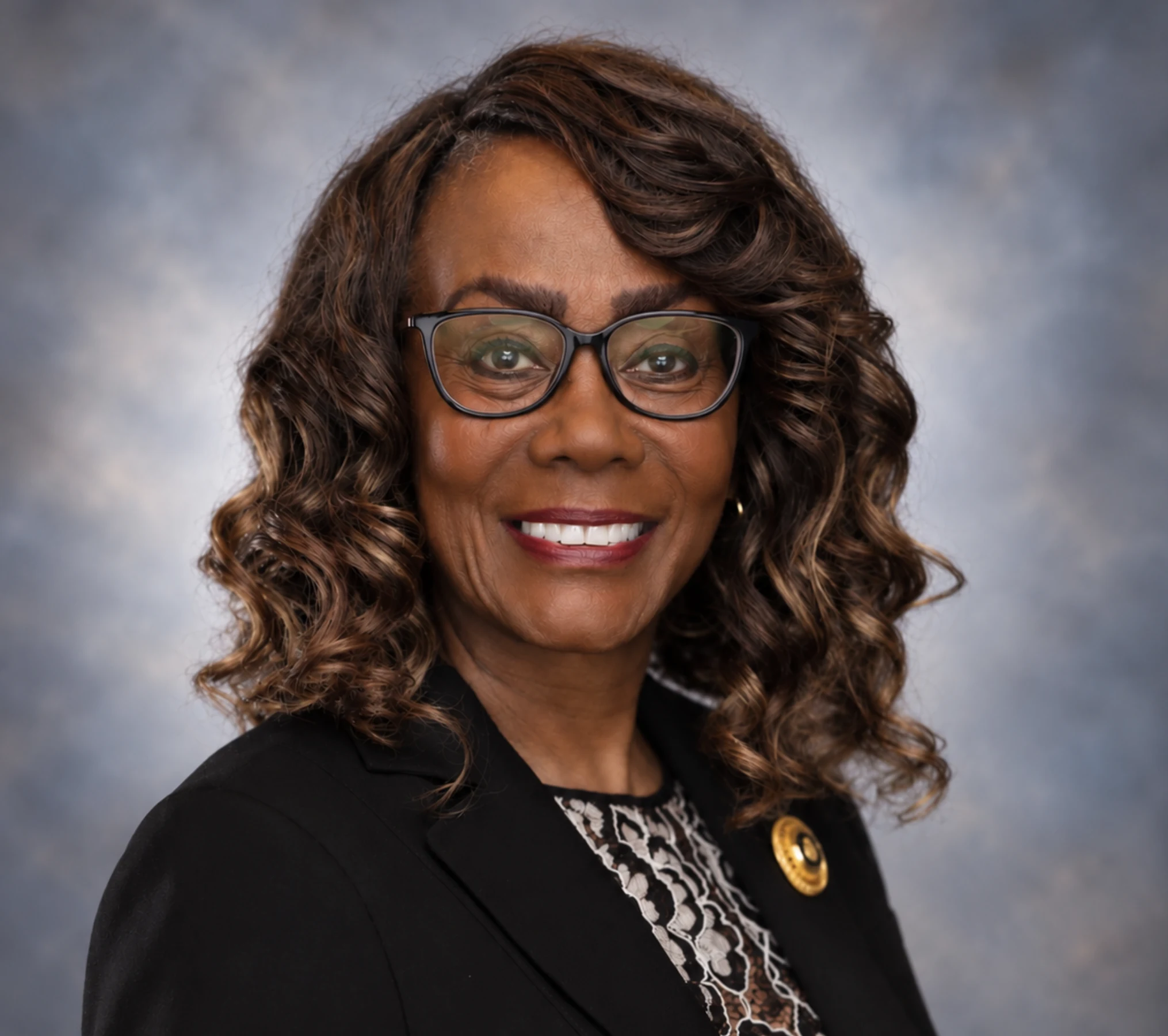 Dianne L Oliphant, smiling in glasses and a black blazer against a softly textured blue-grey studio background.