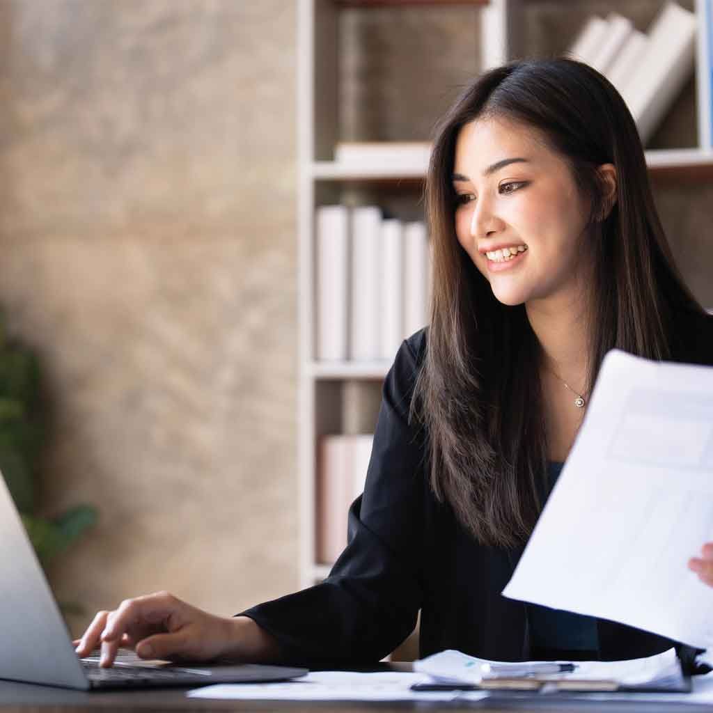 An accountant smiles while working at a laptop, holding documents, in an office.