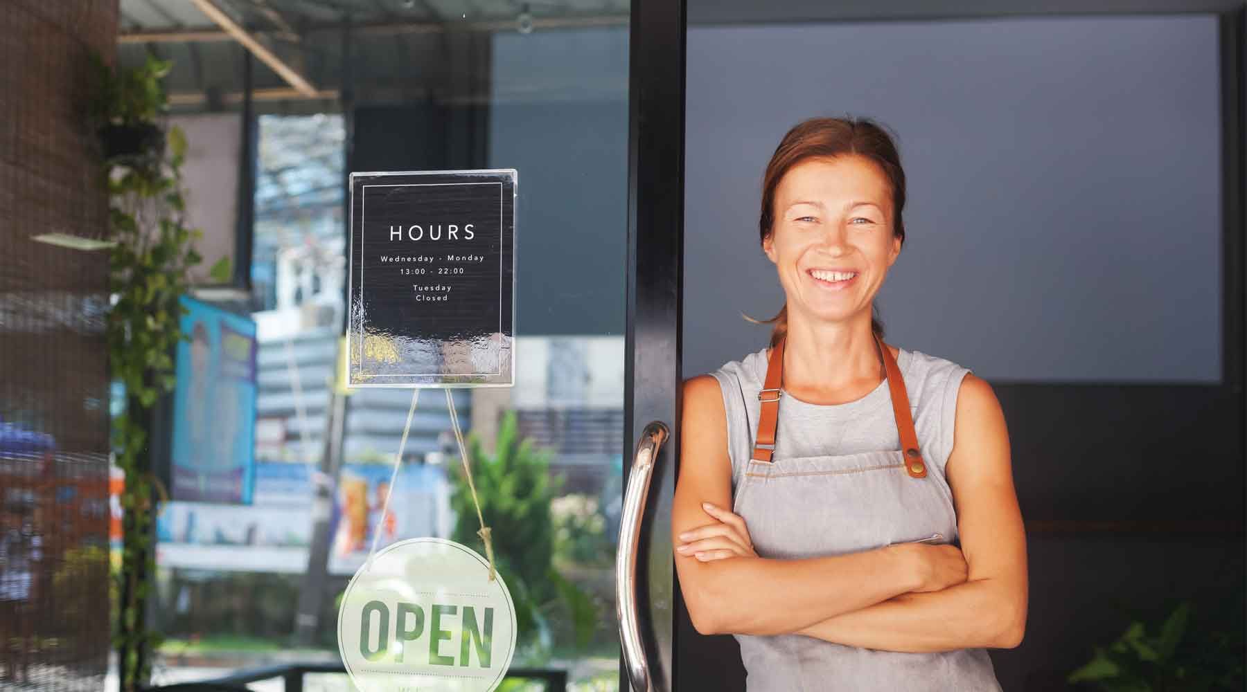 A shop owner in an apron smiles with her arms crossed, standing in her doorway.