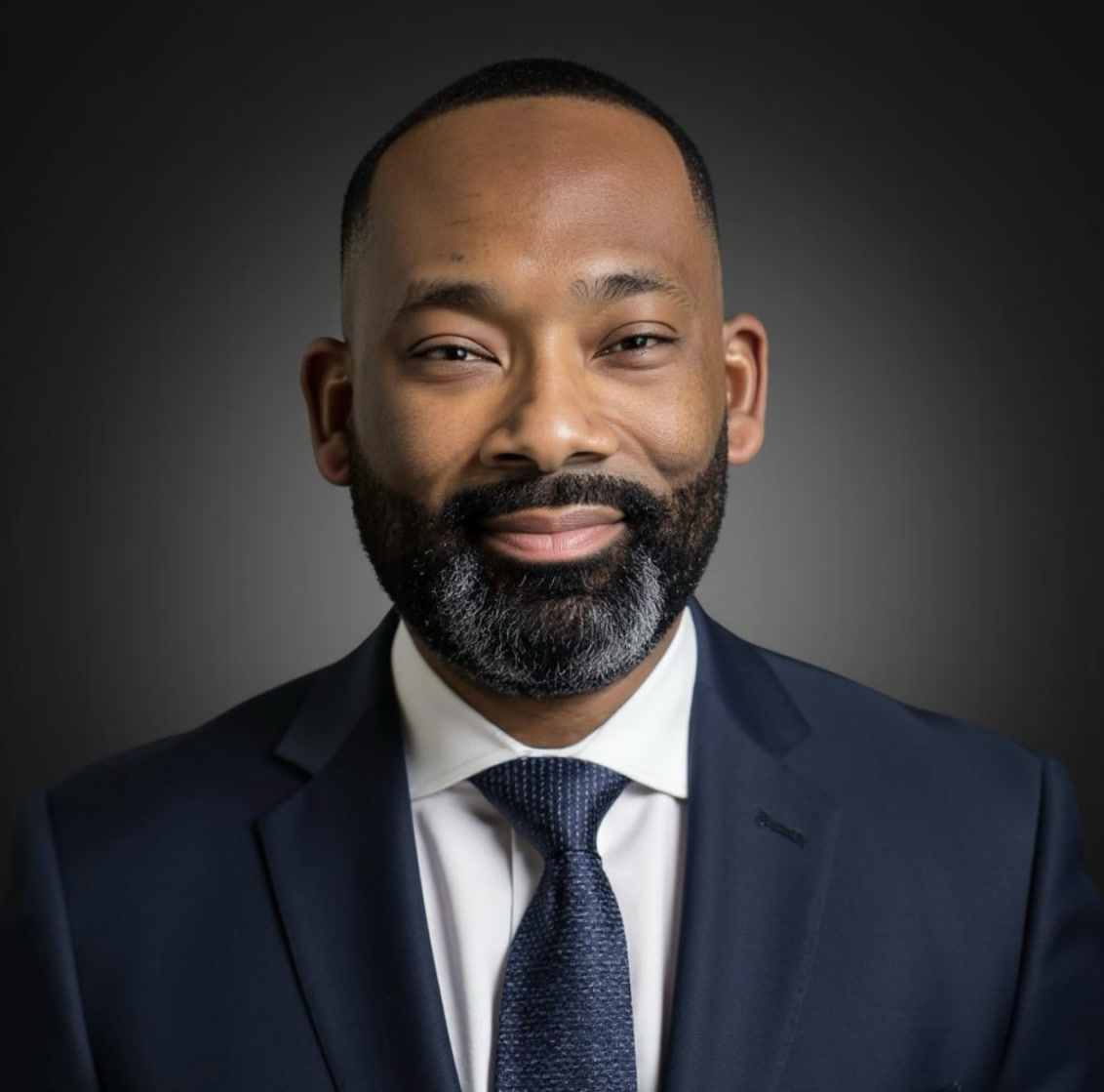 George Reed Jr., in a navy suit and tie, smiling against a dark grey studio background.